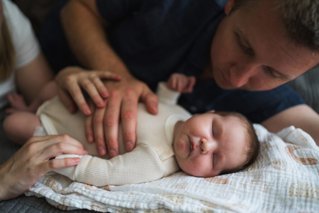 Newborn baby girl laying on bed with father's comforting hand on her during in home portrait session