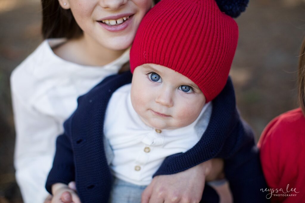 Baby boy in red beanie hat sitting in big sister's lap during portrait session in North Bend, Wa