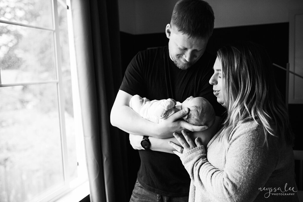Black and white photo of mother and father admiring their newborn baby near the window in the nursery