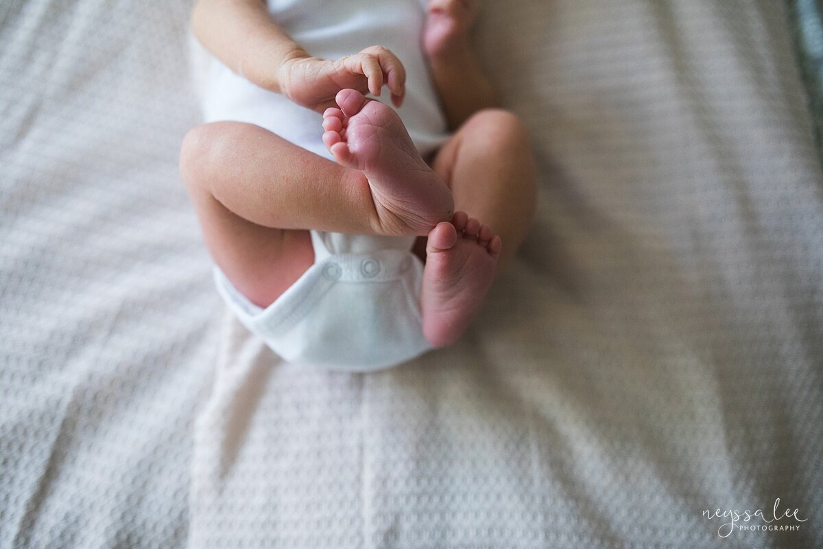lifestyle baby photography of baby in plain white onesie grabbing for her toes on master bed