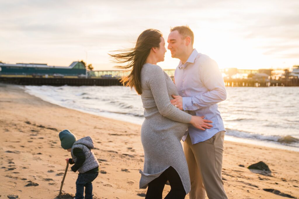 Maternity photos at Brackett's Landing North, photo of boy drawing in sand with a stick behind mother and father