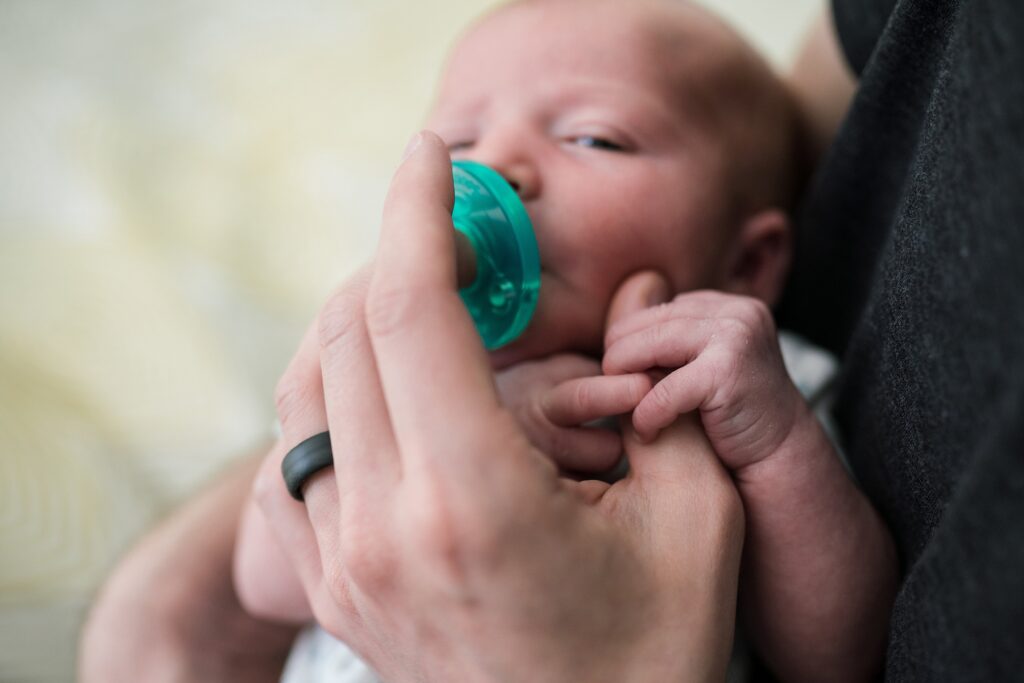 Baby with pacifier holding dad's fingers while waiting for bottle in Seattle newborn portrait session