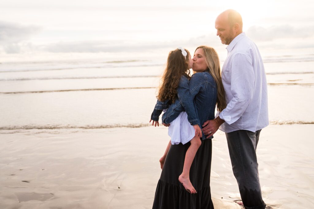 Mother kissing daughter with husband nearby during portrait session at Ocean Shores