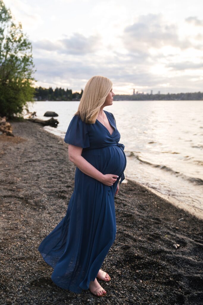 Expectant mother holding baby bump and gazing at the water at sunset during maternity photos in Seattle