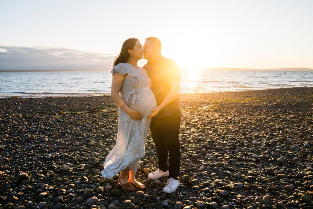 Summer pregnancy picture of husband kissing pregnant wife at sunset at Carkeek Park