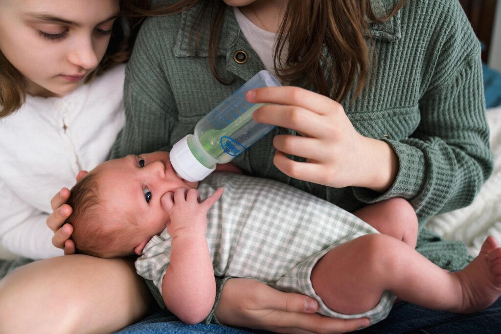 Big sisters giving newborn baby sister a bottle during Seattle area newborn portrait session in their home, when to book newborn photos example