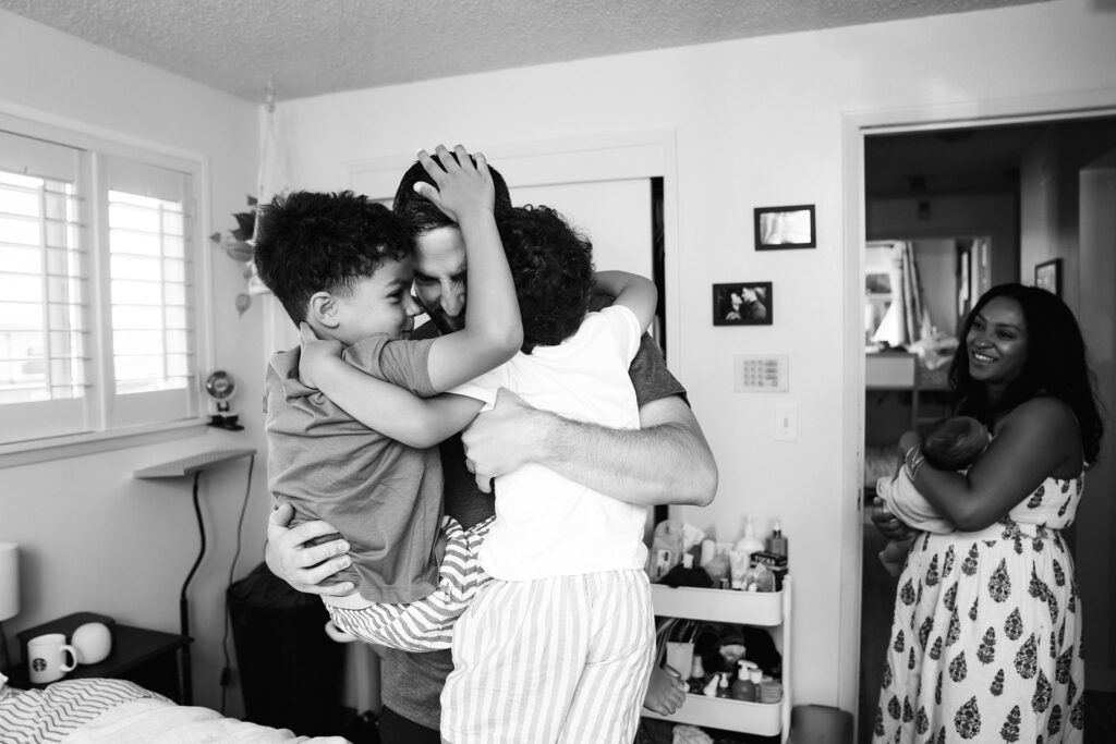 Black and white photo of dad wrestling his boys while mom rocks newborn in background during in home lifestyle newborn photography session by Issaquah photographer